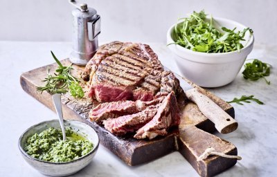 Een grote gegrilde steak ligt op een houten snijplank aangesneden. Er staat een potje homemade rucolapesto langs om meteen bij te scheppen bij je reepje vlees. Er staat ook een metalen pepermolen langs wie het graag wat pittiger wil.