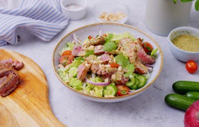 Een heerlijke saladekom is klaargemaakt en staat op het aanrecht nog te pronken. De bowl is rijkelijk gevuld met sla, Chinese kool, komkommerstukjes en tomaat. Hier bovenop liggen de reepjes steak die saignant gebakken zijn. De salade is afgewerkt met de dressing, de overige dressing staat in een potje naast de salade.. 