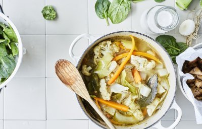 Een lentestoofpotje boordevol groenten, denk maar aan bloemkool, wortel, koolrabi, prei en spinazie. De stoofpot staat op het aanrecht in een grote kookpot, rond de pot liggen nog enkele spinazieblaadjes en een flesje room.