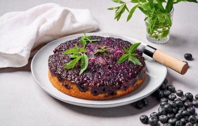 Op tafel staat een soort van tarte tatin, maar eentje met blauwe bessen en cakebeslag. Er ligt een mes langs om aan te snijden en enkele blaadjes verbena. 