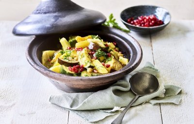 Er staat een tajine op tafel, de tajine heeft een zwarte kleur en is gevuld met stukjes aardappel, courgette, venkel, abrikozen, kikkererwten en granaatappelpitten. 