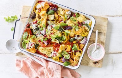 Op een witte tafel staat een houten snijplank met hierop een vierkante ovenschotel. Hij is gevuld met zoete aardappel, courgette, paprika en rode ui. Er is ook Berloumi gebruikt in dit gerechtje. 