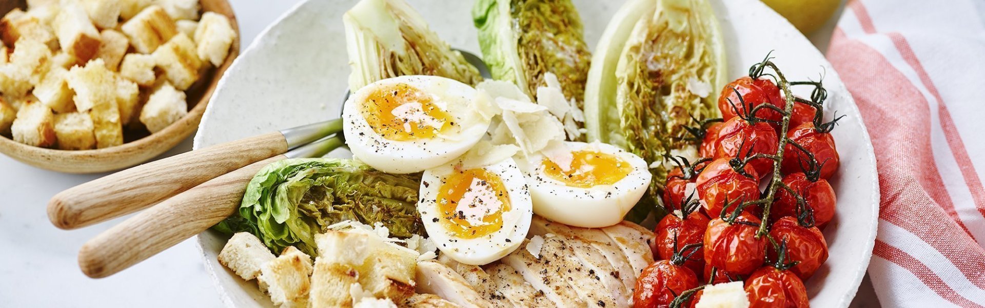 Caesarsalade in een wit diep bord met extra croutons in een houten potje voor de liefhebbers. De dressing op basis van ansjovis staat linksboven. Het bord is rijkelijk gevuld met kip, gegrilde little gem, geroosterde kerstomaatjes en zachtgekookte eitjes.