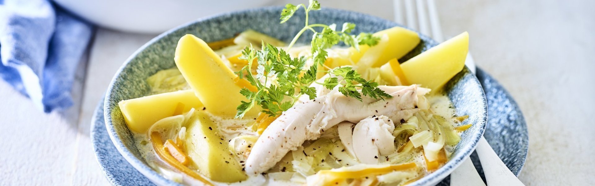 Op een witte houten tafel staat de kookpot vol waterzooi, er is een bordje uitgeschept met hierop de gekookte aardappelpartjes geschikt. Het gerechtje is afgewerkt met een takje verse kervel. 