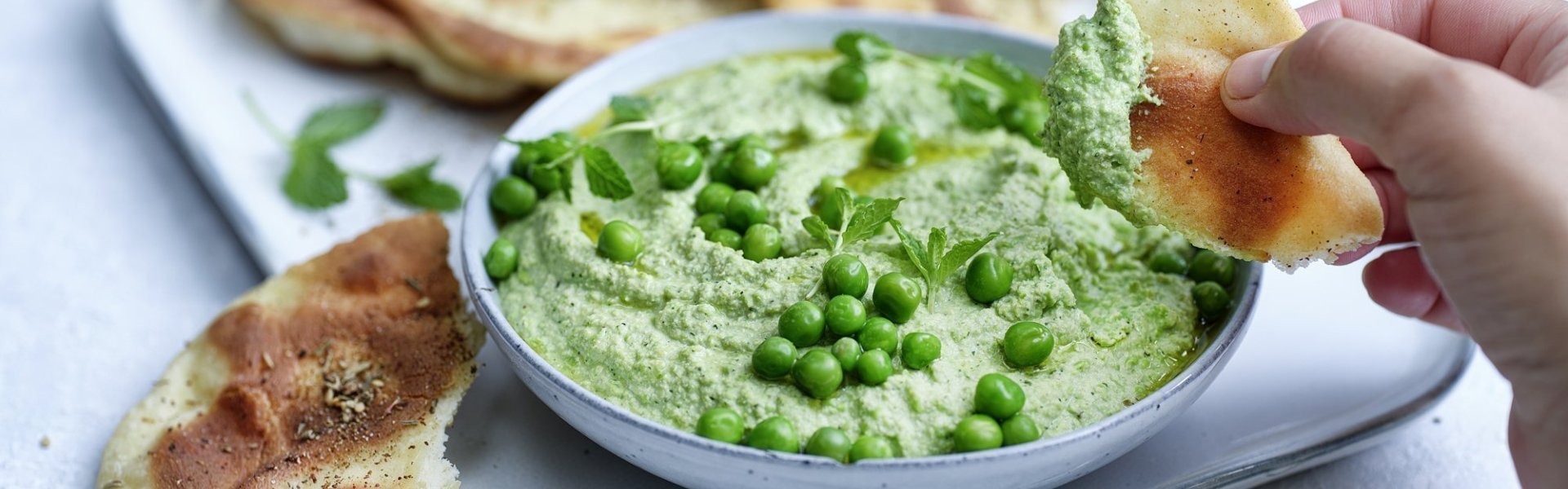 Krokant gebakken platbrood ligt op een serveerschaaltje waarbij een dip van erwten en roomkaas in geserveerd. De dip is gemixt en heeft een mooie groene kleur, om af te werken zijn er nog enkele verse erwtjes bovenop gestrooid. Een hand neem alvast een stukje platbrood om te dippen in gezonde groentedip. 