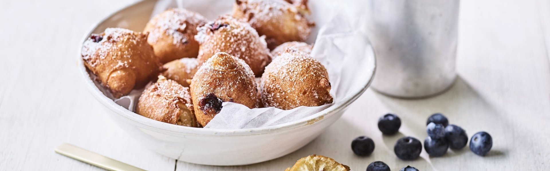 In een diep wit bord ligt een wit boterpapiertje met hierin enkele oliebollen. Er is eentje uitgeprikt en ligt op tafel doormidden gesneden, hier zie je duidelijk de blauwe bessen in zitten. Er is bloemsuiker over de oliebollen gestrooid voor een extra zoete toets. 