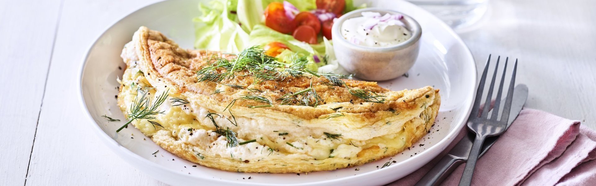 Een heerlijke omelet ligt op een wit bord. Het is een luchtige omelette die we combineren met verse kruiden en kaas. We voegde ook nog wat sla en tomaatjes toe aan het bord voor een extra portie kleur en groenten. 