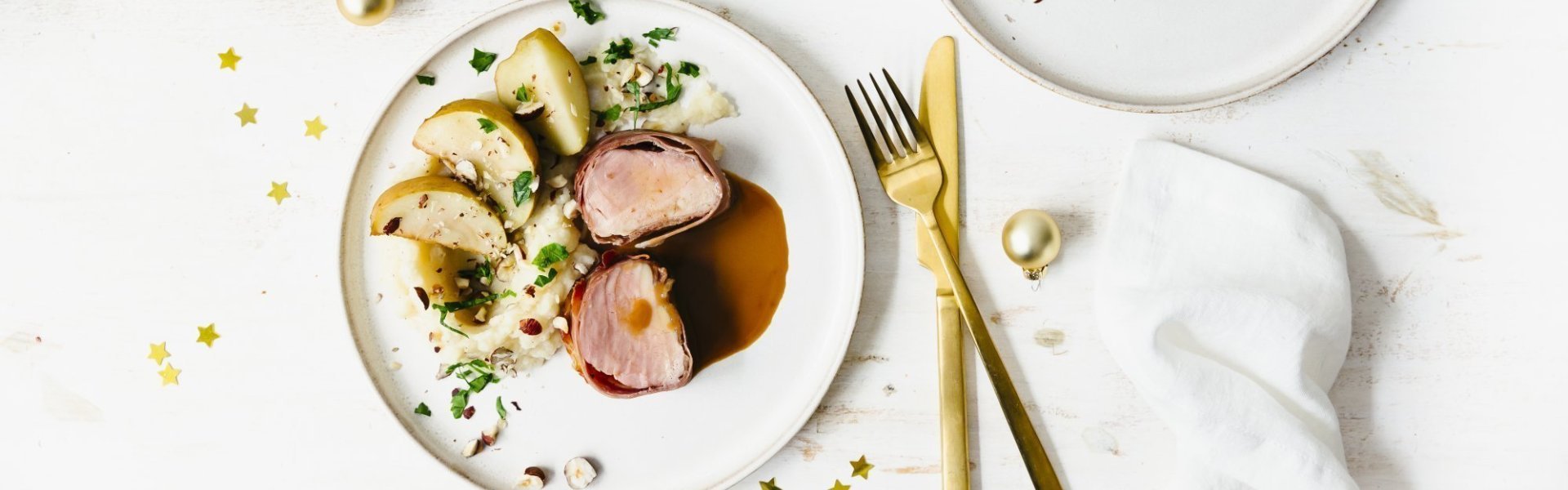 Feestelijk hoofdgerecht van varkenshaasje met een puree van aardpeer en ovenappeltjes. Goud bestek en gouden minikerstballetjes liggen langs het bord en geeft instant een Kerslook.