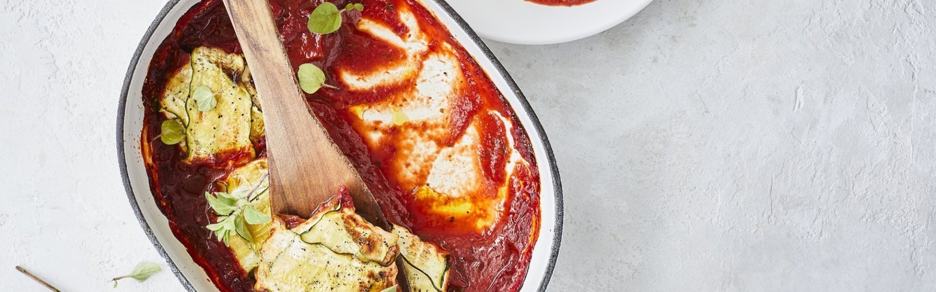 Pakjes van courgette liggen in witte ovenschaal met blauw randje. De tomatensaus ligt onder de ravioli van courgette, waardoor deze goed herkenbaar zijn. Er werd net een portie uitgeschept om meteen te eten. 