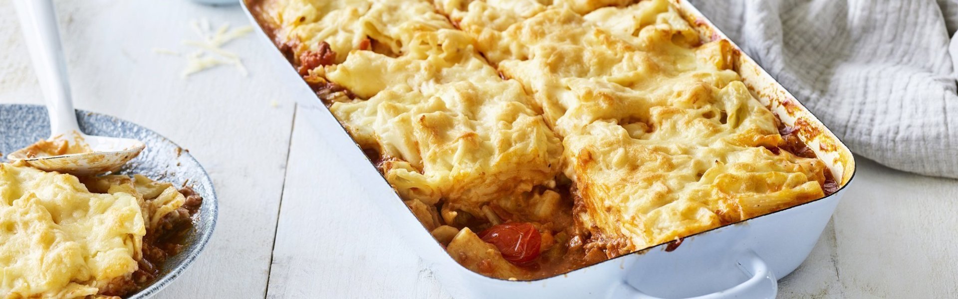 Een lichtblauwe ovenschotel staat op een houten witte tafel. De ovenschotel is gevuld met een mac & cheese én een tomatensaus, waardoor hij eerder weg heeft van een lasagne. Er is al 1 portie uitgeschept en wil al beginnen aan de volgende. Laat het niet koud worden. 