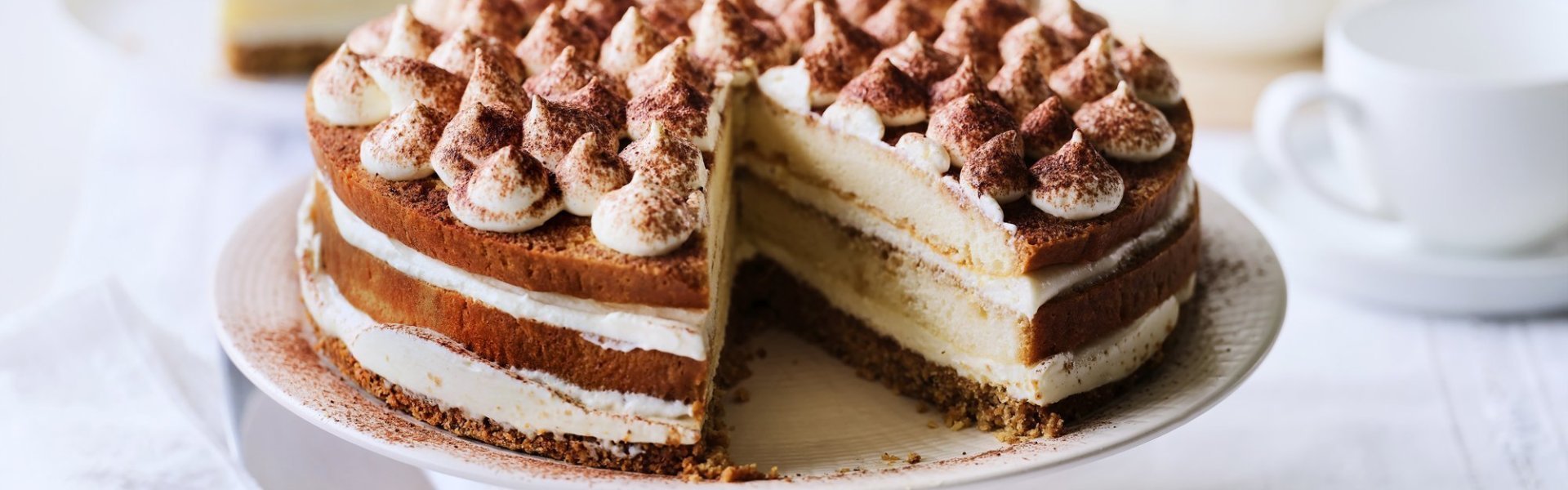 Op tafel staat een prachtige taart, het is een twist op de klassieke tiramisu. Hij staat op een witte taartschaal, en is afgewerkt met toefjes mascarpone en cacaopoeder. In de achtergrond staat een koffiekan en tasjes koffie.