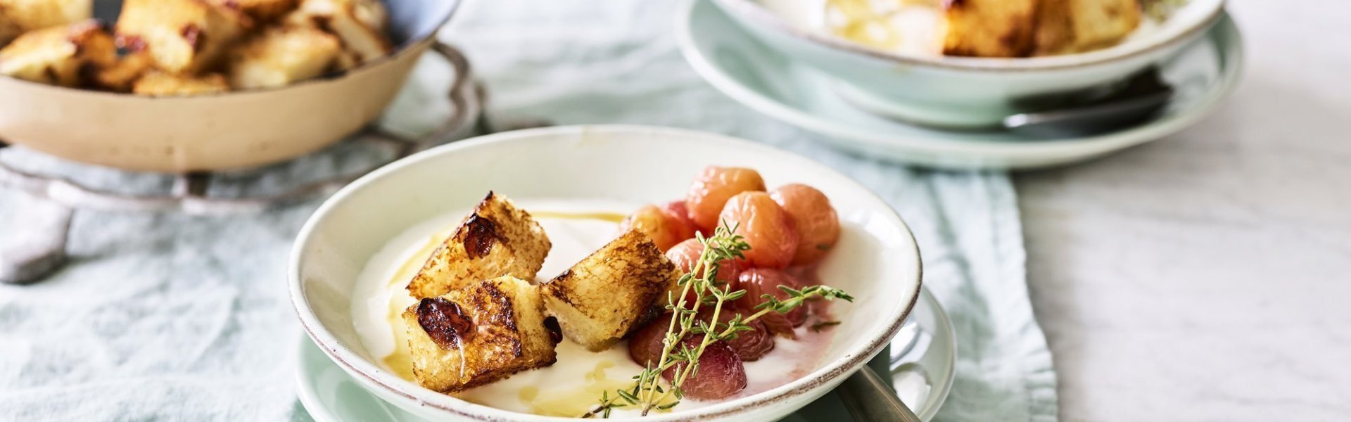 Een heerlijk én makkelijk ontbijtje staat klaar op tafel. De yoghurt is geserveerd in ontbijtkommetjes en afgewerkt met gegrilde druiven en krokante croutons van rozijnenbrood.