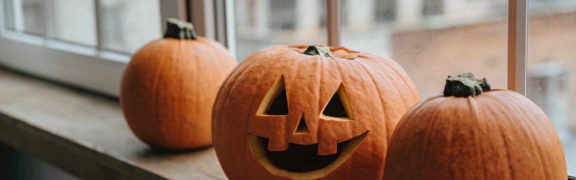 3 pompoenen op een rij, 1 pompoen is uitgehold en heeft een Halloween-gezicht gekregen. Ze staan op de vensterbank, klaar om Halloweenfeest te starten.